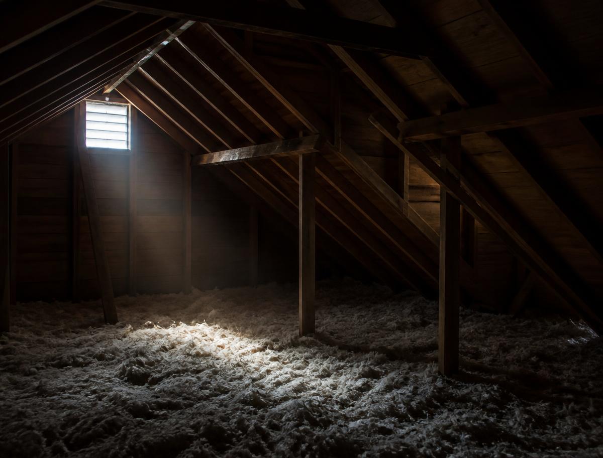 Dim residential attic interior with exposed rafters and loose insulation